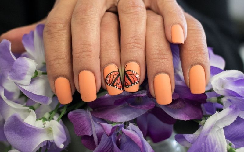 close-up shot captures a girl's hand adorned with butterfly-designed nails painted in a vibrant orange color